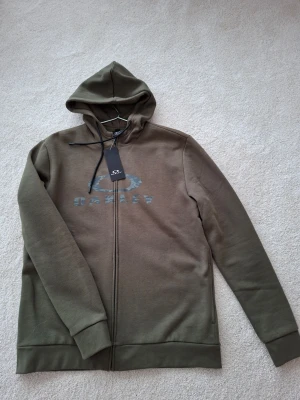 Olivgrön hoodie från Oakley - Snygg olivgrön hoodie från Oakley med dragkedja framtill och stor huva. 
