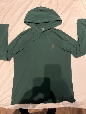 Grön hoodie från Polo Ralph Lauren - Mörkgrön hoodie från Polo Ralph Lauren med klassisk huva och dragsko. Liten röd logga broderad på bröstet. Tillverkad i mjuk bomull som känns skön mot huden. Perfekt för en avslappnad och stilren look.