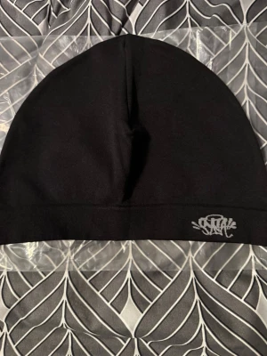 Svart mössa från Synawear - Snygg svart mössa från Synawear med broderad logga framtill. Tillverkad i mjukt tyg som sitter skönt på huvudet. Perfekt för dig som gillar streetwear och vill ha en stilren accessoar med unik touch.