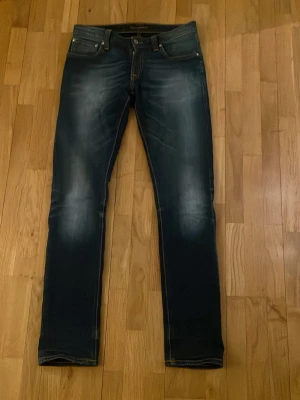 Mörkblå jeans från Nudie Jeans - Säljer ett par mörkblå jeans från Nudie Jeans med snygga slitningar och kontrastsömmar. Klassisk femficksmodell med orange brodyr på bakfickorna och raka ben. Tillverkade i denim med en cool tvättad look.