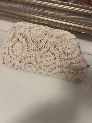 Beige virkad kuvertväska - Säljer en beige kuvertväska med virkat spetsmönster i bomull. Väskan har en mjuk, rektangulär form och stängs med dragkedja. Perfekt för dig som gillar boho-stil och vill ha något unikt till din outfit.