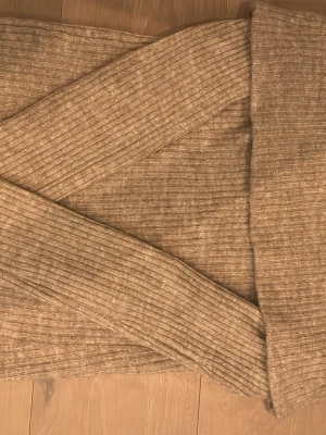 Beige ribbstickad off shoulder-tröja - Säljer en beige ribbstickad tröja med off shoulder-design och långa ärmar. Tröjan har en bred, vikt krage som ger en snygg och avslappnad look. Perfekt för dig som gillar trendiga och bekväma plagg i mjukt material.