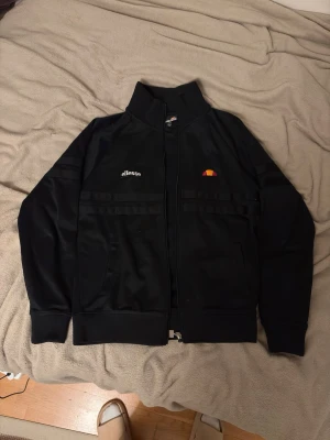 Svart track jacket från Ellesse - Snygg svart track jacket från Ellesse med dragkedja framtill, hög krage och broderad logga på bröstet. Jackan har två fickor fram och diskret text 'Rimini' på ryggen. Perfekt för en sportig och avslappnad stil.