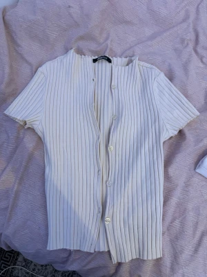 Vit ribbad kortärmad kofta Monki - Säljer en vit ribbad kofta från Monki med korta ärmar och knappar framtill. Koftan har en rak passform och är gjord i ett mjukt, stretchigt material som sitter snyggt på kroppen. Perfekt att styla över ett linne eller t-shirt.