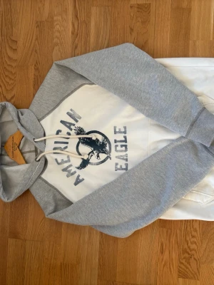 Hoodie american eagle - Snygg hoodie från American Eagle i vitt med ljusgrå ärmar och huva. Stor logga och örntryck framtill. Klassisk känguruficka och dragsko i huvan. Perfekt för en avslappnad och sportig stil. Mjuk bomullsblandning för extra komfort. Vid frågor eller om du är fundersam kontakta🙌