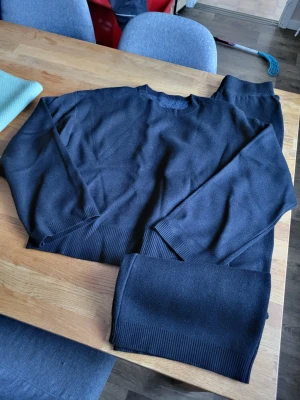 Svart loungewear-set - Mysigt svart loungewear-set med tröja och byxor i mjukt stickat material. Tröjan har rund halsringning och ribbade muddar vid ärmslut och nederkant. Byxorna har elastisk midja och ribbade benslut för extra komfort. Perfekt för chill dagar hemma. Djur finns i hemmet. 