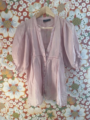 Ljusrosa blus med puffärm från Lindex - Söt ljusrosa blus från Lindex med puffiga trekvartsärmar och dekorativa knappar framtill. Blusen har v-ringning och fina spetsdetaljer längs sömmarna. Tillverkad i ett tunt, luftigt tyg som ger en avslappnad och romantisk känsla. Tappat en knapp.