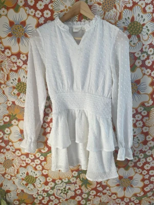 Vit volangblus med smockdetalj - Söt vit blus med lång ärm, smock i midjan och volang nedtill. Blusen har diskret prickstruktur i tyget, liten volangkrage och v-ringning. Perfekt för dig som gillar romantisk och trendig stil. Tillverkad i ett lätt och luftigt material. Liten fläck vid kragen.