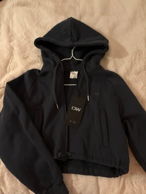 Mörkgrön cropped hoodie från ICANIWILL - Snygg mörkgrön cropped hoodie från ICANIWILL med dragsko i huvan och elastisk nederkant. Hoodien har långa ärmar, två fickor framtill och diskret ICW-logga på bröstet. Tillverkad i mjukt material som känns skönt mot huden.  Aldrig använd. Prislappen kvar. Orginalpris: 699kr