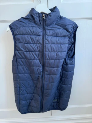 Mörkblå dunväst - Snygg mörkblå dunväst från Lindbergh med dragkedja framtill. Västen har quiltad design och två sidofickor med dragkedja. Perfekt för lager-på-lager och ger en sportig look. Materialet är lätt och smidigt, vilket gör den enkel att bära.