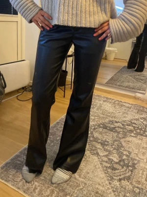 Svarta utsvängda läderbyxor i fuskskinn - Snygga svarta läderbyxor i fuskskinn med utsvängda ben. Byxorna har hög midja och en glansig finish som ger en trendig look. Perfekta för dig som vill sticka ut lite extra och gillar en edgy stil.