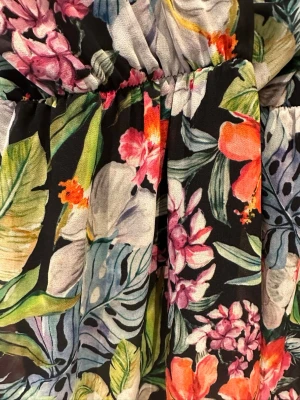 Långklänning med färgglada blommor - Säljer en långklänning med svart botten och färgglatt blommönster i rosa, orange, vitt och grönt. Klänningen har en luftig passform och är perfekt för sommarens varma dagar. Materialet känns lätt och mjukt mot huden.