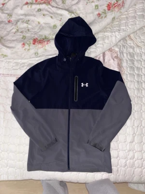 Blå och grå vindjacka från Under Armour - Snygg tvåfärgad vindjacka från Under Armour i marinblått och grått. Jackan har huva med dragsko, hel dragkedja framtill och en vertikal bröstficka med dragkedja. Perfekt för blåsiga dagar och sportiga outfits. Nypris 1,250, säljs inte längre 