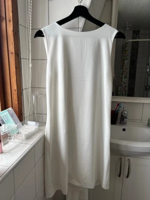 Vit ärmlös klänning med rosett - Stilren vit klänning i kort modell med djup v-ringning och breda axelband. Klänningen har en stor dekorativ rosett i midjan och är tillverkad i ett mjukt tyg som ger ett elegant fall. Perfekt för dig som vill ha en stundtklänning eller lite finare vit. Ny med prislapp. Originalpris - 1300kr