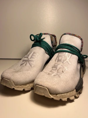 Adidas Pharrell Hu NMD vita sneakers - Unika Adidas Pharrell Hu NMD sneakers i vitt mesh med gröna skosnören och brokigt mönstrad kant runt plösen. Skorna har asiatiska tecken på ovandelen och en chunky sula i beige. Perfekt för dig som gillar statement sneakers och streetwear.
