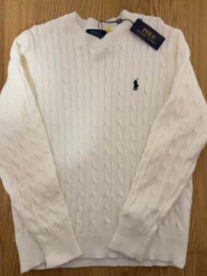 Vit Stickad  Polo Ralph Lauren Tröja - Kabelstickad vit tröja från Polo Ralph Lauren med klassisk rund halsringning och broderad marinblå logga på bröstet. Tröjan har långa ärmar och ribbade muddar. Perfekt för dig som gillar stilrena och tidlösa plagg.
