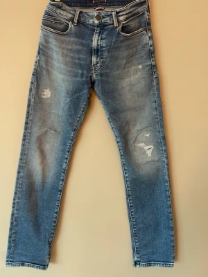 Blå jeans från Tommy Hilfiger - Snygga blå jeans från Tommy Hilfiger med slitningar på båda benen och klassisk femficksdesign. Jeansen har raka ben och tydliga sömmar, samt diskret logga på bakfickan. Perfekta för en avslappnad och trendig look.