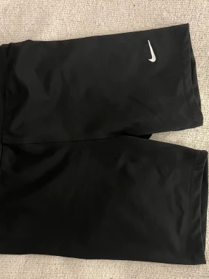 Svarta träningsshorts från Nike - Säljer ett par svarta cykel/tränings shorts från Nike med den klassiska vita swoosh-loggan på låret. Shortsen har en tight passform och är tillverkade i ett stretchigt syntetmaterial som passar perfekt för träning eller sportiga outfits.