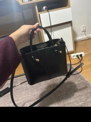 Svart handväska med gulddetaljer - Snygg svart handväska i slätt konstläder med både handtag och justerbar axelrem. Väskan har guldfärgade metalldetaljer och dragkedja upptill samt en innerficka för småsaker. Perfekt storlek för det viktigaste och stilren design.