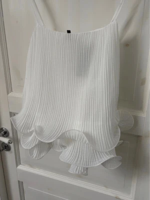 drömmig vit topp med volang  - en drömmig vit topp i plisserat material med tunna axelband och volangkant nertill ☁️ lätt och luftig med fint fall, perfekt till sommaren. aldrig använd, prislapp kvar. nypris 400 kr. i mycket fint skick ✨