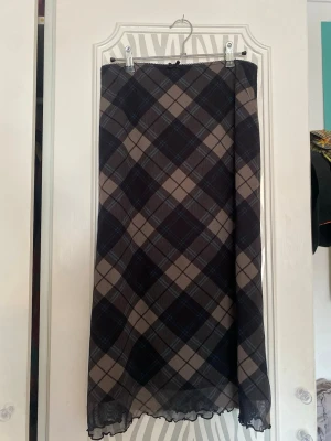 Rutig långkjol med rosett - Snygg långkjol med rutigt mönster i brun, grått och beige. Kjolen har en liten söt rosett upptill och vågig kant nertill. Från urban outfitterd, har ett litet hål där lappen suttit innan (visas på bild) syns knappt annars. Tror storleken är S