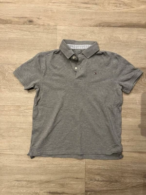 Grå pikétröja Tommy Hilfiger - Klassisk grå pikétröja från Tommy Hilfiger med korta ärmar och liten logga på bröstet. Tillverkad i mjuk bomull och har krage med knappar framtill. Perfekt för en clean och enkel stil.
