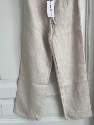 Beiga linnebyxor Design by Si x Matilde W - Snygga beiga linnebyxor från Design by Si x Matilde W. Byxorna har rak passform, resår i midjan med snörning och är tillverkade i ett luftigt linnematerial. Perfekta för en chill och stilren look.