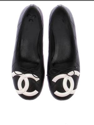 Chanel Ballerines noires logo Black - Säljer ett par svarta ballerinaskor från Chanel i quiltat läder med stora vita CC-loggor framtill. Skorna har rund tå och små rosetter på ovansidan. Sula med Chanel-tryck i rött. Perfekta för dig som gillar klassisk och lyxig stil.