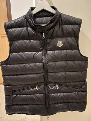 Svart dunväst från Moncler - Snygg svart dunväst från Moncler med quiltad design och hög krage. Västen har två dragkedjeförsedda fickor framtill och en tydlig Moncler-logga på bröstet. Perfekt för lager-på-lager och håller dig varm med stil.