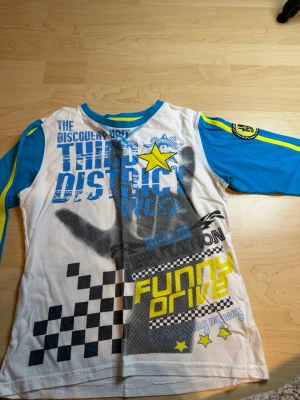 Färgglad långärmad t-shirt WeTweens - Säljer en cool långärmad t-shirt från WeTweens by KappAhl med blå ärmar, vit bas och tryck i blått, svart och gult. T-shirten har grafiska detaljer, stjärnor och text som ger en streetstyle-vibe. Perfekt för dig som gillar färg och mönster!