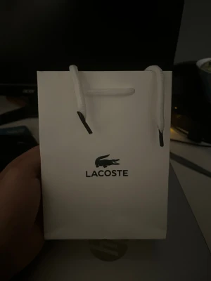 Vit presentpåse och ask från Lacoste - Säljer en stilren vit presentpåse och matchande ask från Lacoste. Båda har det klassiska gröna krokodilloggan och är tillverkade i kraftigt papper med struktur. Perfekt för att ge bort accessoarer eller små presenter.