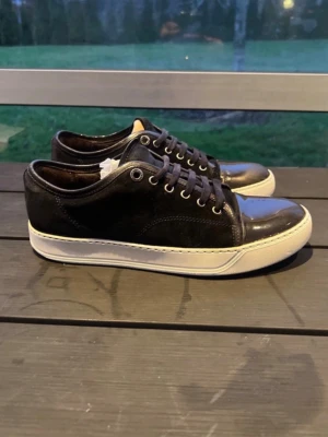 Byter Lanvin  - Snygga svarta sneakers med lågt skaft och vit sula. Skorna har svarta skosnören, metallfärgade öljetter och en stilren design. Ovandelen är i en mix av läder och mocka vilket ger en schysst kontrast. Perfekta för dig som gillar en clean och klassisk look.