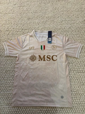 Vit Napoli matchtröja EA7 - Snygg vit Napoli matchtröja från EA7 med guldfärgade detaljer och MSC-logga på bröstet. Tröjan har italienska flaggan och klubbmärke, samt diskret mönster i tyget. Tillverkad i lätt och ventilerande funktionsmaterial, perfekt för träning eller match.