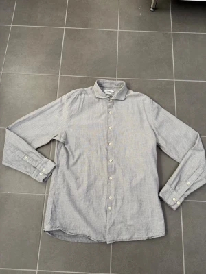 Grå skjorta från J.Lindberg i bomull - Stilren grå skjorta från Arket med diskret mönster. Skjortan har klassisk krage, långa ärmar och vita knappar framtill. Tillverkad i mjuk bomull som känns skön mot huden. Perfekt för en clean och enkel look.