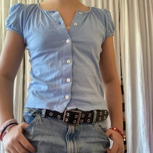 Ljusblå kortärmad blus med knappar - Supersöt ljusblå blus med korta puffärmar och vita knappar framtill. Blusen är figurnära i modellen. Perfekt att styla med jeans eller kjol för en fräsch look. Märket är franskt och är ett mycket bra kvalitetsmärke