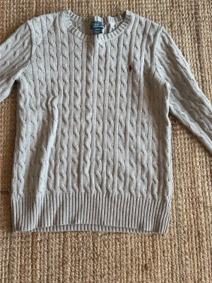 Ljusgrå kabelstickad tröja Polo Ralph Lauren - Kabelstickad tröja i ljusgrått från Polo Ralph Lauren med rund halsringning och ribbade muddar. Klassisk röd logga broderad på bröstet. Perfekt till jeans eller kjol för en clean och stilren look. Priset kan diskuteras 