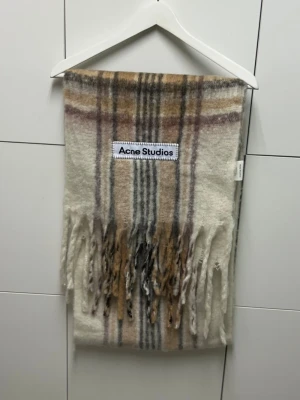 Acne Studios Halsduk - Mysig halsduk från Acne Studios i ullblandning med rutigt mönster i beige, grått och brunt. Stor logotyp framtill och långa fransar i ändarna. Perfekt accessoar för att lyfta din outfit med en trendig touch.