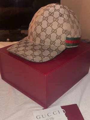 äkta Gucci beige keps  storlek s - Säljer en beige keps från Gucci med det klassiska GG-monogrammet över hela kepsen. På sidan finns den ikoniska gröna och röda randen. Kepsen har böjd skärm och justerbar rem bak. Materialet är canvas med exklusiv känsla. Perfekt för dig som vill ha något lyxigt och trendigt. kvitto lådan finns