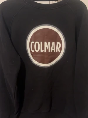 Svart sweatshirt från Colmar - Säljer en svart sweatshirt från Colmar Originals med stort tryckt logotypmotiv i vitt och brunt på bröstet. Tröjan har rund halsringning och är tillverkad i mjukt bomullsmaterial. Perfekt för en avslappnad och stilren look.