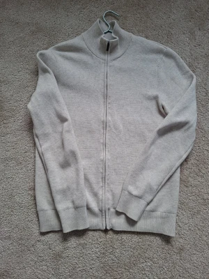 Beige ziptröja från Bläck - Snygg beige ziptröja från Bläck med hög krage och ribbade muddar. Tröjan är tillverkad i mjukt stickat material och har en stilren, minimalistisk look.