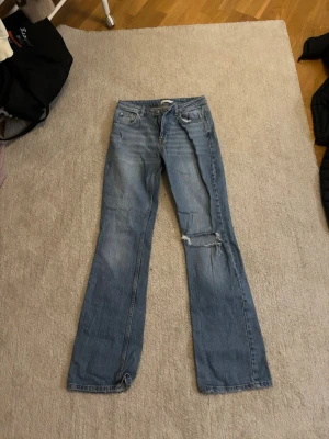 Blå bootcut jeans med slitning - Säljer ett par ljusblå bootcut jeans med slitning på ena benet och klassisk femficksdesign. Jeansen har en avslappnad passform och är tillverkade i denim. Perfekta för en casual look med lite retrovibe.
