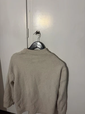 Beige stickad tröja med rund hals - Mysig beige stickad tröja med rund hals och långa ärmar. Tröjan har en enkel, klassisk design med ribbstickade muddar vid hals, ärmslut och nederkant. Perfekt för lager på lager och en avslappnad stil.