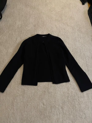 Svart kofta  - Stilren svart kofta  med rund halsringning och en knapp framtill. Koftan har lång ärm och rak passform, perfekt att slänga över en topp. Materialet ser ut att vara mjukt och tunt, troligen bomull eller en bomullsblandning.