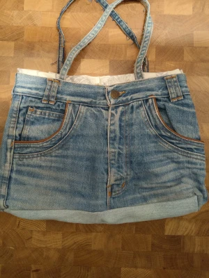 Handväska i denim med jeanslook - Unik handväska sydd av återvunna blå jeans med klassiska fickor fram och bak, bälteshällor och kontrastsömmar. Väskan har två axelband i samma denimtyg och en cool, avslappnad streetstil. Perfekt för dig som gillar kreativ och personlig stil.