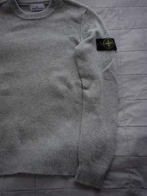 Stone Island Knit Grey  - Säljer en ljusgrå stickad tröja från Stone Island med klassisk rund hals och ribbade muddar. Tröjan har den ikoniska Stone Island-loggan på vänster ärm. Perfekt för dig som gillar stilrena och tidlösa plagg.