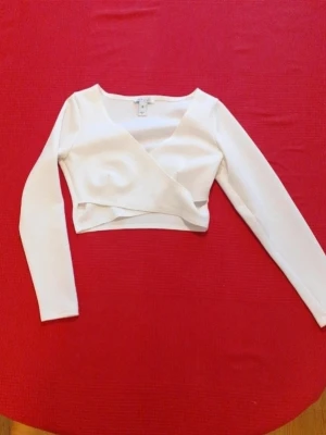 Vit omlottopp från H&M - Snygg vit croppad omlottopp från NLY Trend med lång ärm och v-ringad hals. Tillverkad i mjuk bomullsmix som känns skön mot huden. Perfekt att styla med höga jeans eller kjol för en trendig look.