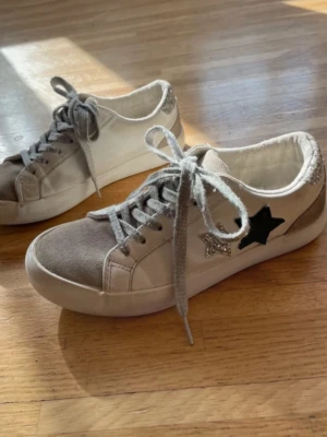 Vita sneakers med stjärndetaljer - Snygga vita sneakers med grå mockadetaljer på tån och silverfärgade glitterstjärnor på sidan. Skorna har även en svart stjärna och silverglittrig häl. Klassisk låg modell med grå skosnören och vit sula. Ny pris på skorna är 500kr