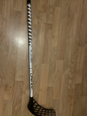 Unihoc innebandyklubba svart/vit - Säljer en innebandyklubba från Unihoc i svart och vitt med texten 'PLAYER' på skaftet. Klubban har ett svart blad med rutmönster och ett vitt skaft med svarta detaljer. Perfekt för dig som gillar innebandy och vill ha en snygg och funktionell klubba.