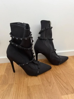 Svarta stövletter med nitar och klack - Säljer ett par svarta stövletter med spetsig tå och höga smala klackar. Skorna har dekorativa remmar med svarta nitar runt skaftet och foten. Dragkedja på insidan för enkel påtagning. Snygg och edgy design som sticker ut. Storlek 7, passar 38. 11 cm klack.