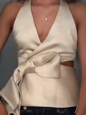 champangefärgad omlottopp med rosett - Snygg champangefärgad/beige omlottopp med stor rosettdetalj i midjan och djup v-ringning. Toppen är ärmlös och har en elegant, figurnära passform som framhäver midjan. Perfekt för dig som vill ha en stilren men ändå unik look.